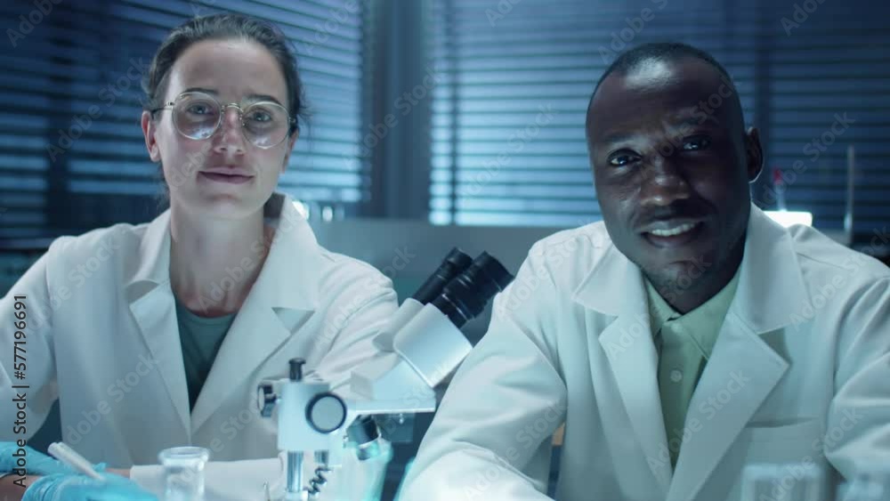 Portrait of diverse male and female scientists sitting at desk with microscope and posing together for camera while working in laboratory Portrait of diverse male and female scientists sitting at desk with microscope and posing together for camera while working in laboratory
