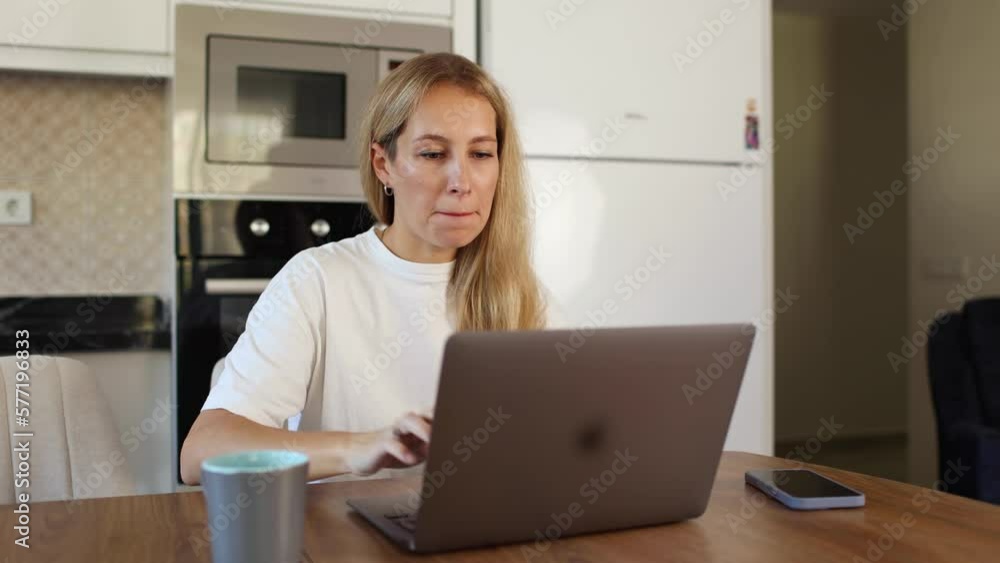 Young business woman in white t-shirt holding cup of tee while working with her laptop sitting at table Young business woman in white t-shirt holding cup of tee while working with her laptop sitting at table
