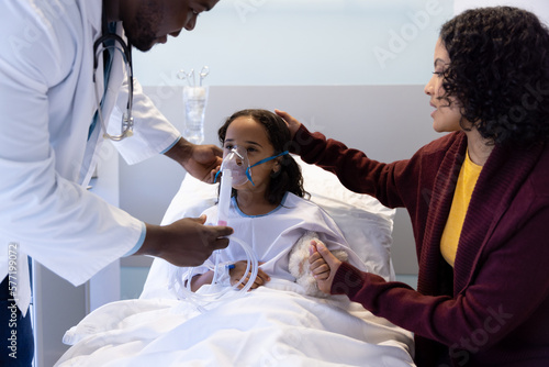 Diverse male doctor examining sick daughter with mother with oxygen mask and teddy bear on bed photo