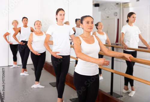 Group of women engaged in classical ballet in a dance studio stand holding onto a barre in a ballet stance, perfoming the ..1st position with their hands and the 3rd position with their feet