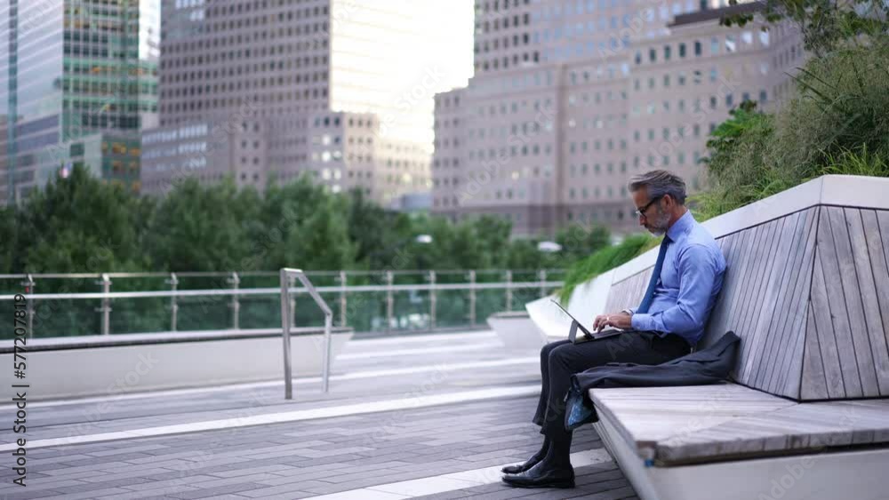 Full length view, Successful Mature businessman in blue shirt and suit sitting on bench while typing text for email on touch pad. Caucasian entrepreneur in stylish eye glasses using tablet for work Full length view, Successful Mature businessman in blue shirt and suit sitting on bench while typing text for email on touch pad. Caucasian entrepreneur in stylish eye glasses using tablet for work