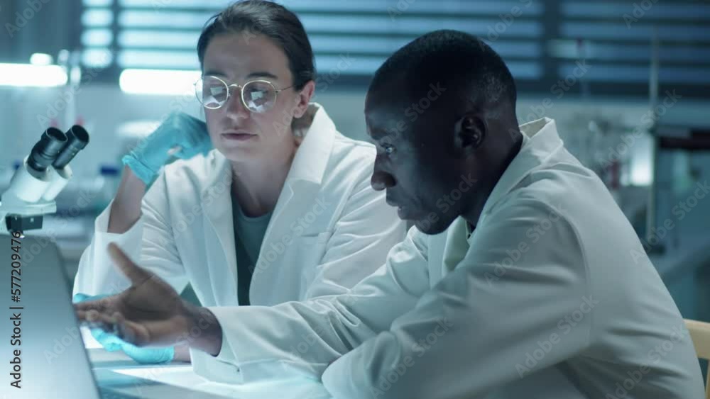 Medium shot of two multiethnic scientists discussing something on laptop while working together in laboratory Medium shot of two multiethnic scientists discussing something on laptop while working together in laboratory