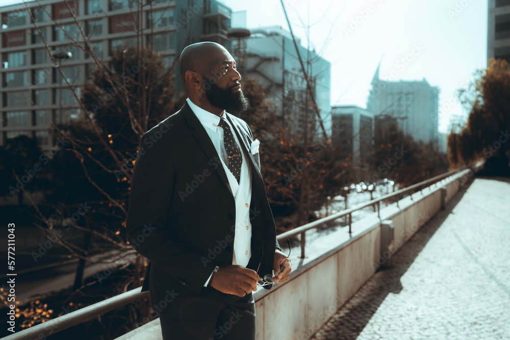 A bald, well-dressed black man leans against an iron railing on the sidewalk, clutching a pair of sunglasses; dressed to impress in a sharp suit and tie, and sports a long, well-groomed beard A bald, well-dressed black man leans against an iron railing on the sidewalk, clutching a pair of sunglasses; dressed to impress in a sharp suit and tie, and sports a long, well-groomed beard