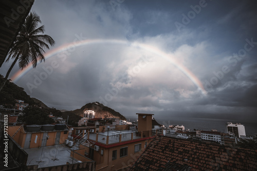 From a favela, a wide-angle low-key view of a balcony facing a backdrop of dark, moody skies; a beautiful rainbow shines through the clouds, adding a burst of color and hope to the scene; photo