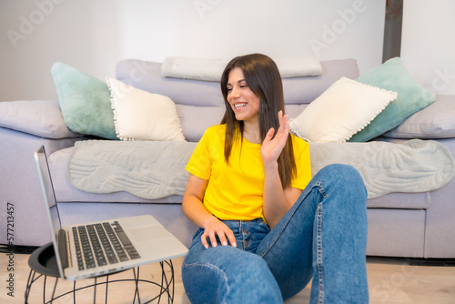 Woman with a computer sitting on a sofa, millennial, social media, waving on a video call © unai