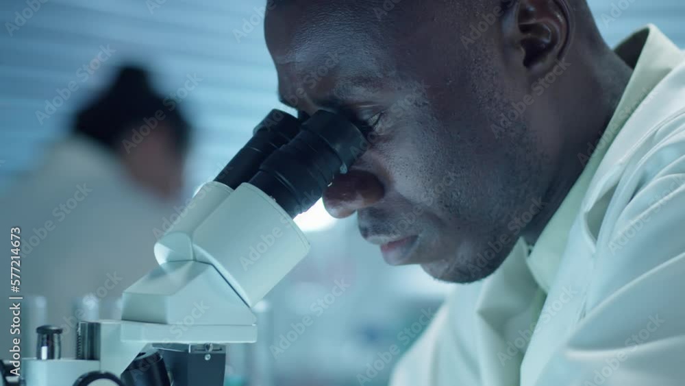 Close up arc shot of African American male scientist in lab coat examining sample through microscope while doing research in laboratory Close up arc shot of African American male scientist in lab coat examining sample through microscope while doing research in laboratory