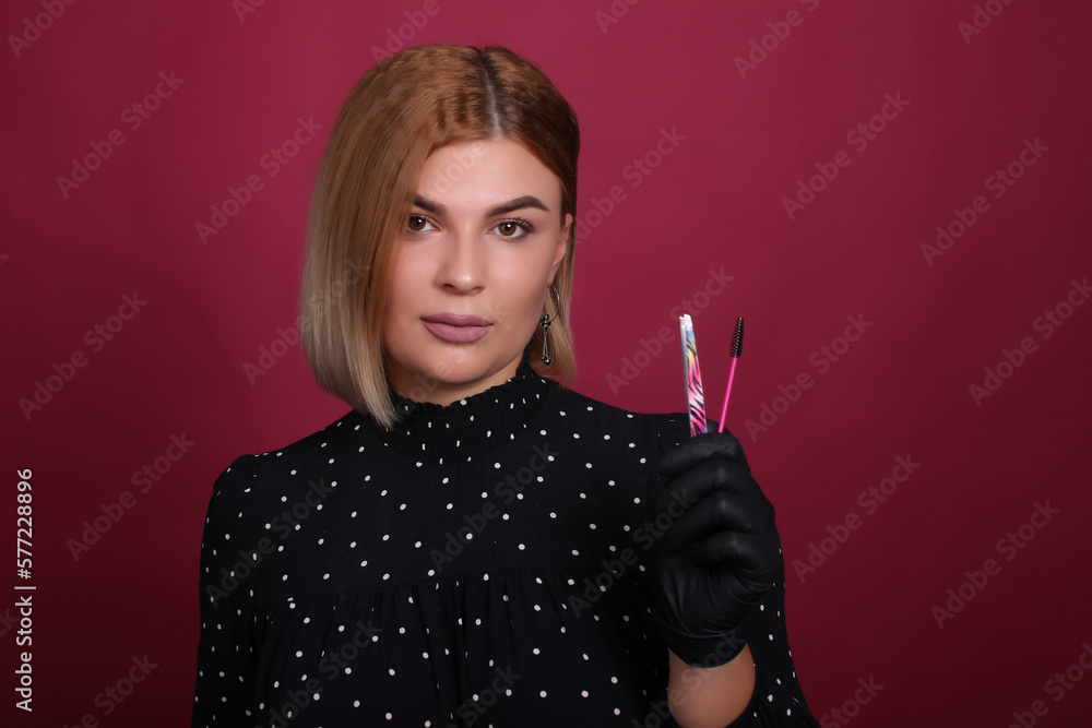Obraz premium portrait of a natural blonde girl of an eyelash master with blonde hair in a black dress in black disposable gloves in a studio room, with makeup and hairstyle, with mascara devices in her hands