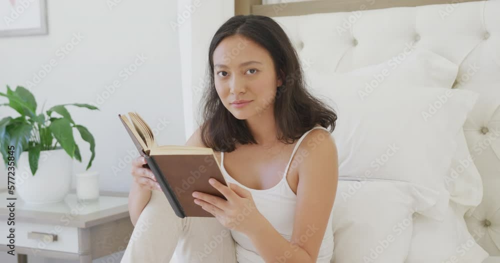 Portrait of happy asian woman sitting on bed and reading book, in slow motion Portrait of happy asian woman sitting on bed and reading book, in slow motion