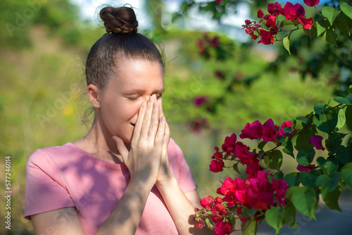 Portrait of beautiful young allergic woman is suffering from pollen allergy or cold on natural flower flowering tree background at spring or sunny summer day sneezes, blowing her runny nose rubs eyes