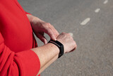 Elderly female hands in red sportswear checking time, text message or pulse using digital smartwatch against background of road. Concept of modern technologies and healthy lifestyle.
