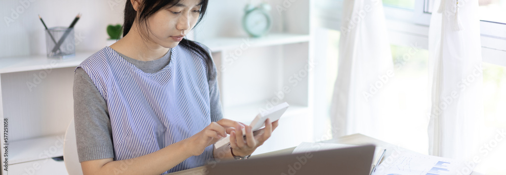 Asian woman working at home, Press the calculator to calculate the income - expenses of the house and use the laptop to record the information and check the accuracy, Home lifestyle. Asian woman working at home, Press the calculator to calculate the income - expenses of the house and use the laptop to record the information and check the accuracy, Home lifestyle.