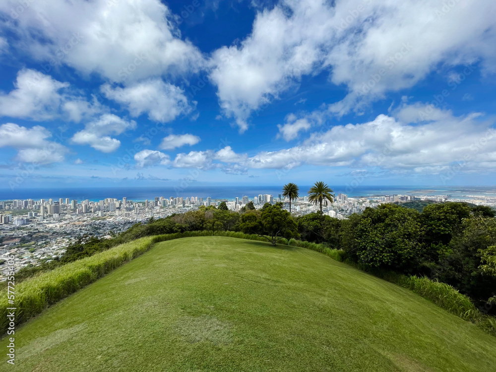 Green grass mountain hill with cloudy blue skies on a sunny day with buildings and city in the bakground in Oahu Hawaii