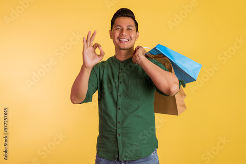 handsome asian man standing with oke hand gesture and holding the shopping bags on his shoulder on orange isolated background photo