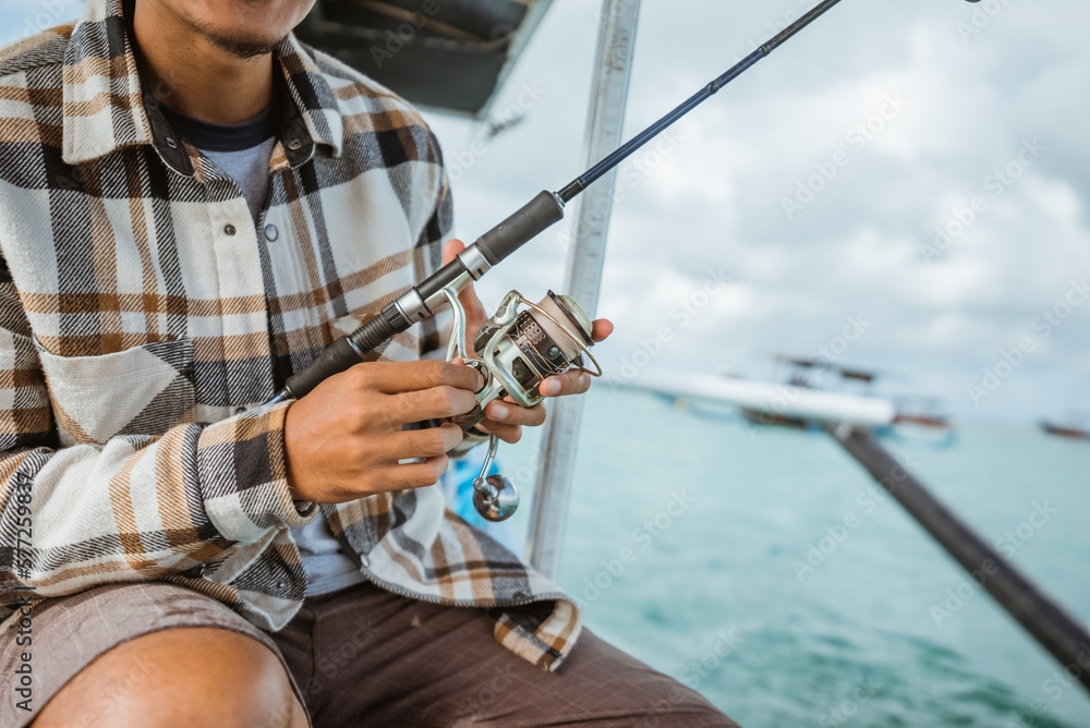 Obraz premium close up of angler's hand checking reel while holding fishing rod on small fishing boat