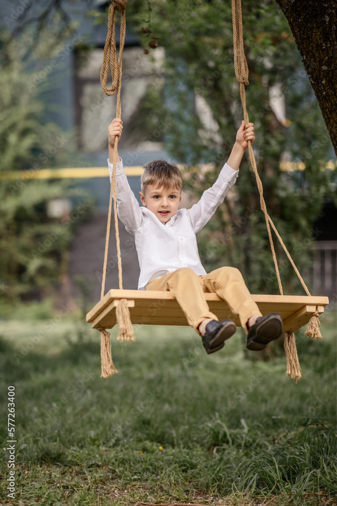 A little boy 6-7 years old in a white shirt is resting, swinging on a swing in the spring in the garden of a country house. the concept of village life, healthy lifestyle