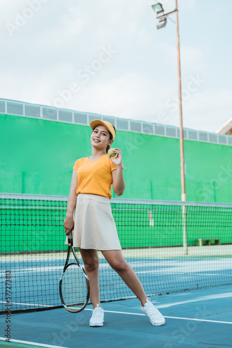 asian woman in tennis sport suit and hat holding racket and ball against net background