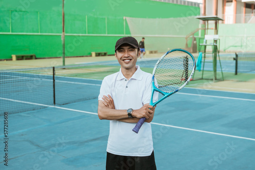Smiling male athlete with crossed hands holding a racket against a tennis court in the background © Odua Images