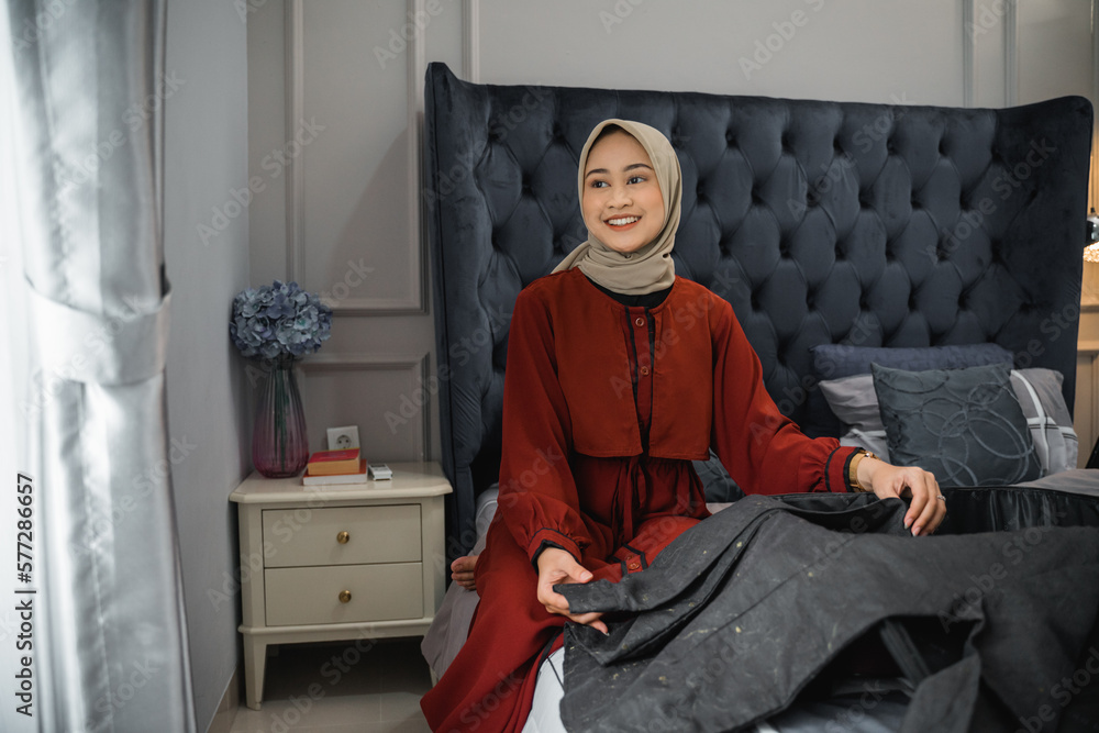 Beautiful woman in hijab smiles looking at the window while folding clothes sitting on the bed