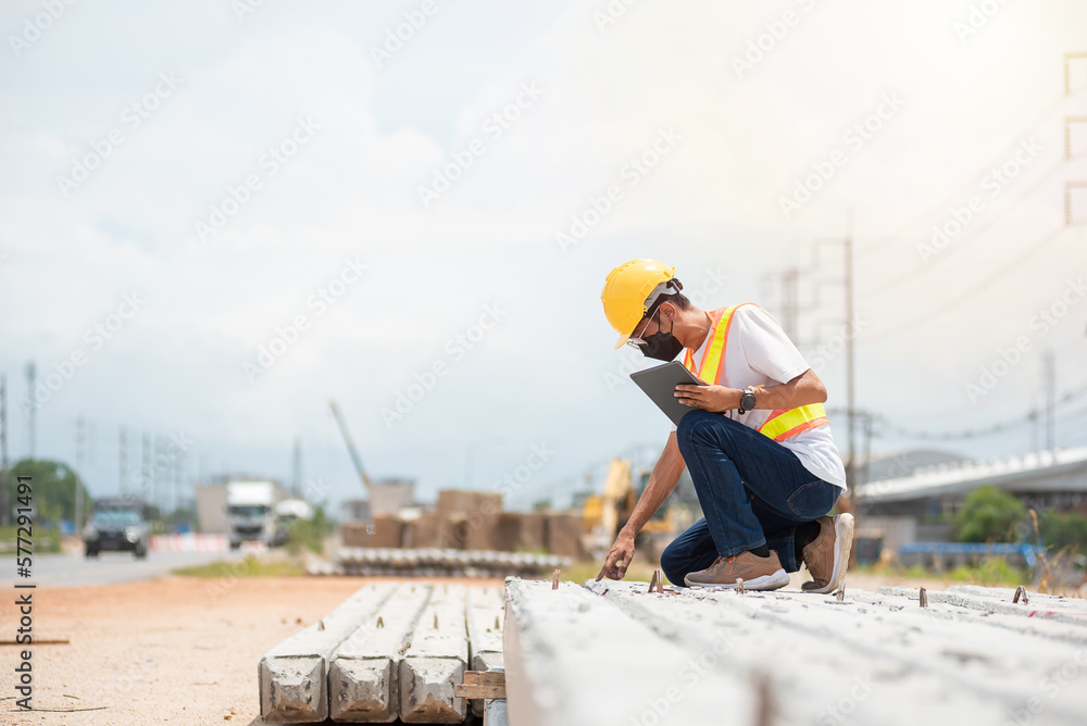 Asian engineer wearing hard hat and safety vest sitting pointing finger to the construction while holding tablet in construction site. Asian engineer wearing hard hat and safety vest sitting pointing finger to the construction while holding tablet in construction site.