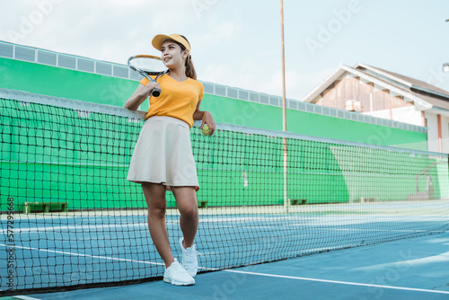 woman in hat and sportswear holding racket and ball leaning on net in tennis court
