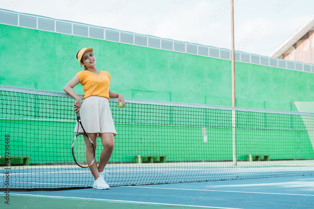woman in tennis sportswear holding racket and ball leaning on net in tennis court woman in tennis sportswear holding racket and ball leaning on net in tennis court