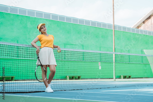woman in tennis sportswear holding racket and ball leaning on net in tennis court