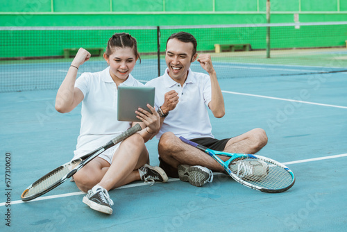 excited doubles tennis players sitting looking at pad screen together on tennis court
