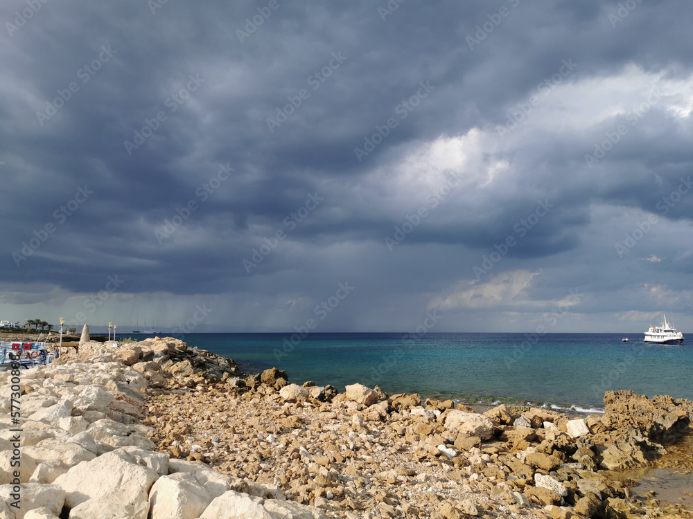 The stone coast of the Mediterranean Sea against the backdrop of the sea with a walking white ship and a dramatic sky.