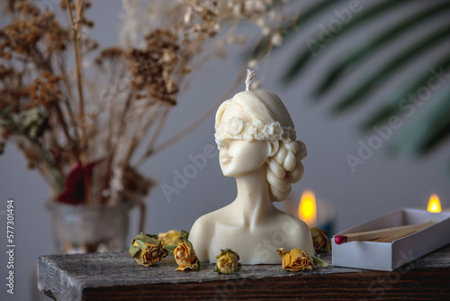 Beautiful feminine handmade candle in the shape of a female body on a wooden stand. There are matches and yellow rose buds nearby