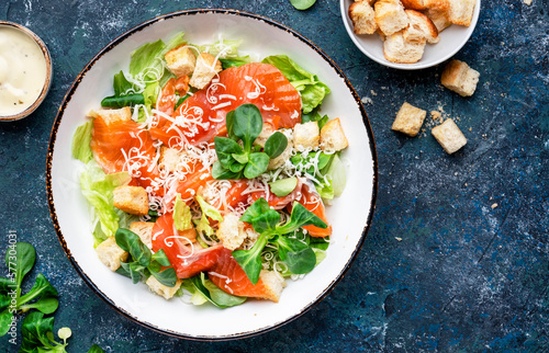 Caesar salad with salted salmon, iceberg and lamb lettuce, croutons, parmesan cheese with caesar dressing. Blue stone table background, top view