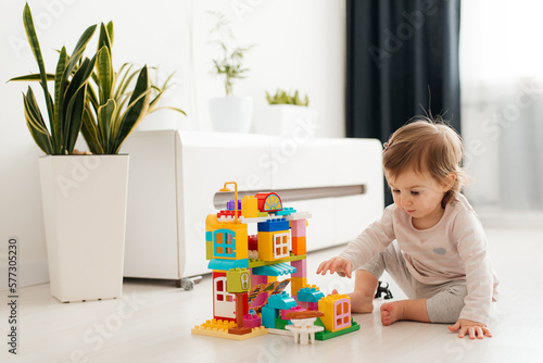 Baby girl playing indoors sitting on floor in playroom at home with constructor. Educational game for baby and toddler. Children build toy house. Creative