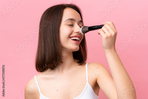 Young Ukrainian woman isolated on pink background holding makeup brush and whit happy expression