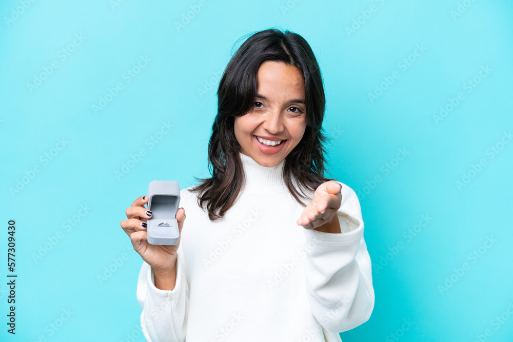 Young hispanic woman holding a engagement ring isolated on blue background shaking hands for closing a good deal Young hispanic woman holding a engagement ring isolated on blue background shaking hands for closing a good deal