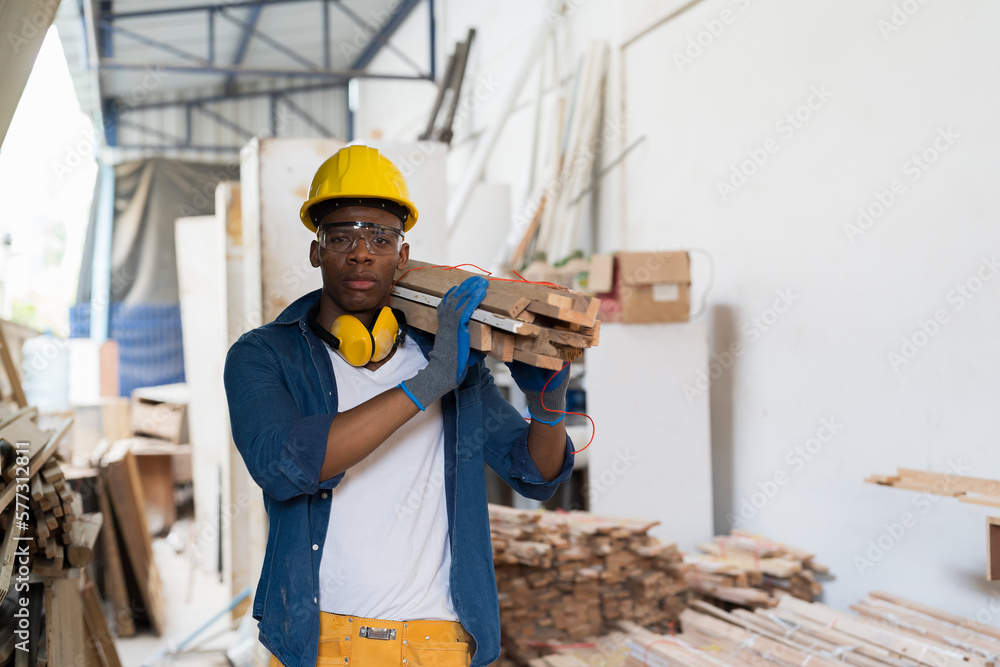 Male carpenter carrying piece of wood on his shoulder at wood processing plants. Black male carpenter working at wood workshop. Young man carpenter at furniture workshop Male carpenter carrying piece of wood on his shoulder at wood processing plants. Black male carpenter working at wood workshop. Young man carpenter at furniture workshop