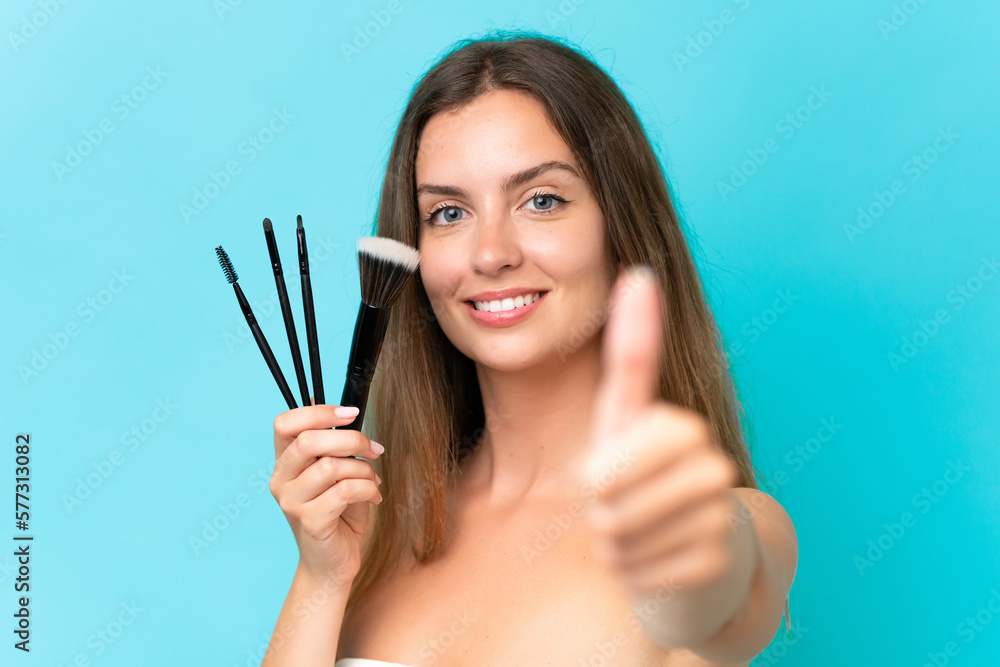 Young caucasian woman isolated on blue background holding makeup brush and with thumb up Young caucasian woman isolated on blue background holding makeup brush and with thumb up