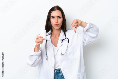 Young caucasian woman isolated on white background wearing a doctor gown and holding pills while doing bad signal