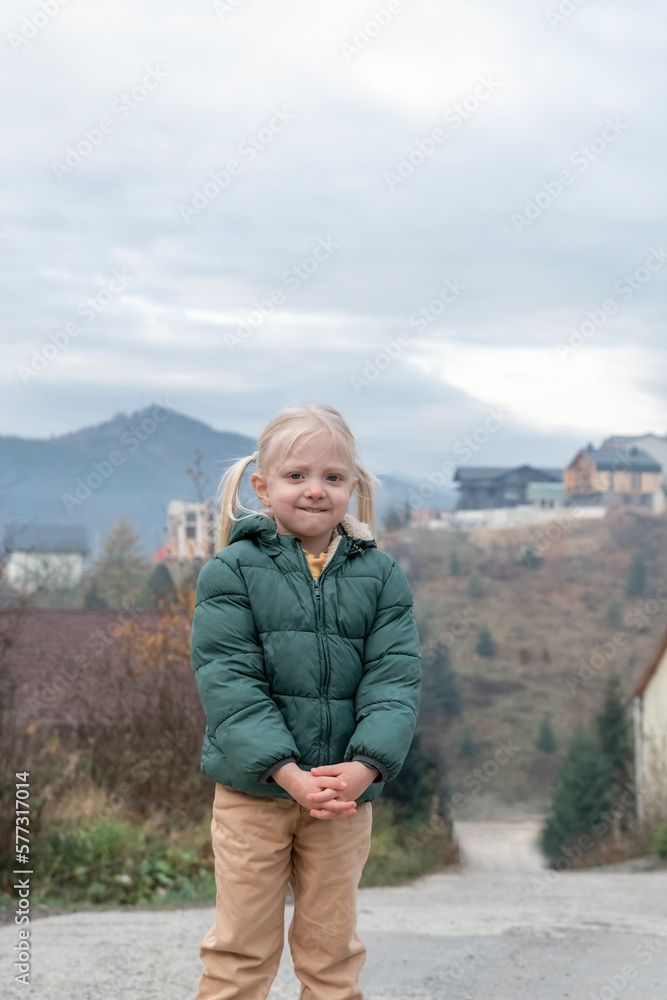 Cute fair-haired girl with two ponytails wears warm green autumn jacket, mountain background. Vertical frame.