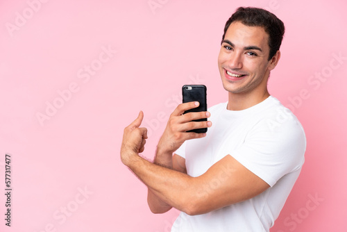 Young caucasian man isolated on pink background using mobile phone and pointing back © luismolinero