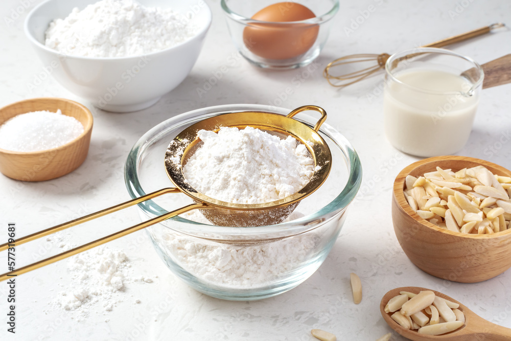 Sifting flour with gold sieve in glass bowl with baking ingredients on marble kitchen table