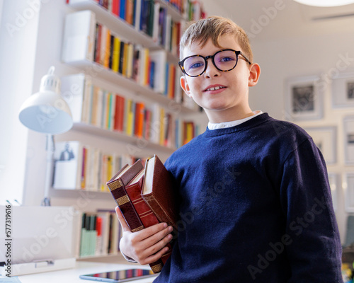 Bambino biondo con gli occhiali vestito sta in piedi con dei libri in mano dentro lo studio photo