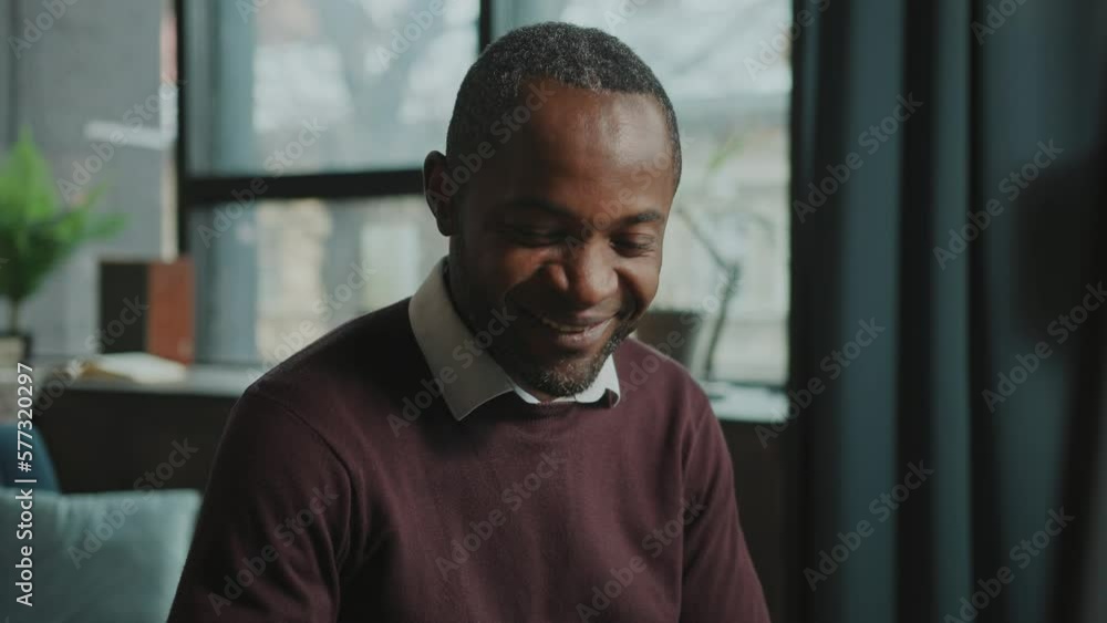 Portrait of thoughtful African man sitting at computer focused on work looking at screen and reading message while working from home. Smiling businessman enjoying job. Indoors. Blurred background of Portrait of thoughtful African man sitting at computer focused on work looking at screen and reading message while working from home. Smiling businessman enjoying job. Indoors. Blurred background of