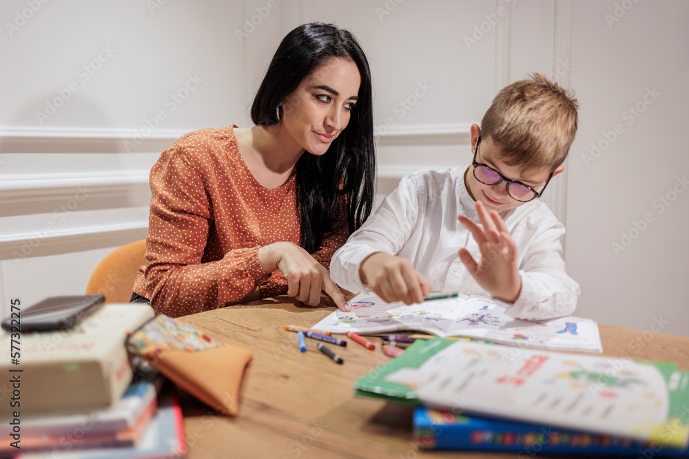 Una mamma con i capelli scuri aiuta nel fare i compiti il proprio bambino biondo con gli occhiali , all'interno di  in un ambiente elegante.