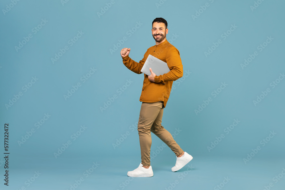 Full length of happy man holding laptop pc computer and doing winner gesture clenching fist isolated on blue background