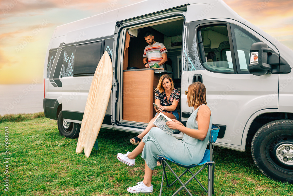 Group of friends traveling in a camper van. Two women talking in front of  camper van while young man cooks foto de Stock | Adobe Stock