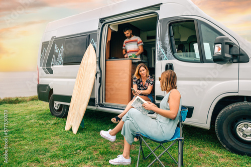Group of friends traveling in a camper van. Two women talking in front of camper van while young man cooks