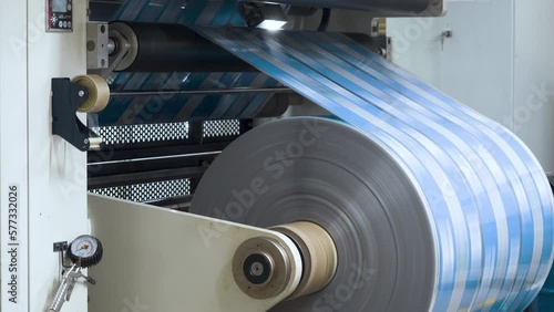 Flexo printing. Printing house. Large roll rotates rapidly. Film spins between shafts. Roller machine prints. Anilox shafts