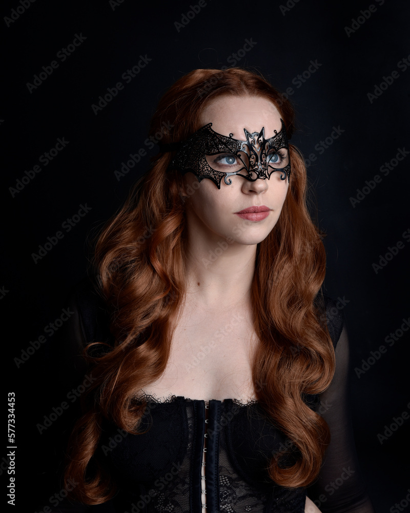 Masked close up portrait of beautiful woman with long red hair wearing sheer corset top, and black bat wing mask. Isolated on dark studio background with. Moody silhouette lighting. Masked close up portrait of beautiful woman with long red hair wearing sheer corset top, and black bat wing mask. Isolated on dark studio background with. Moody silhouette lighting.