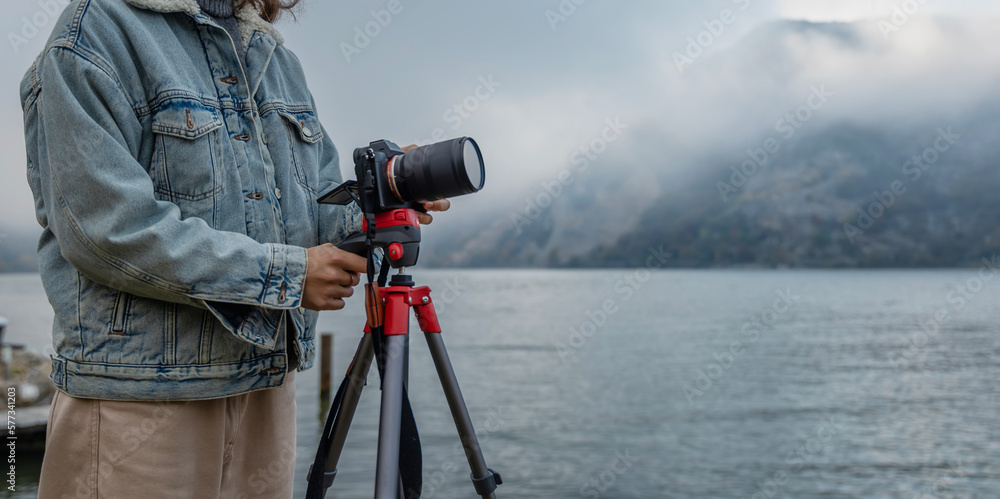 Close-up shot of a photo camera on a tripod and the hands of a woman photographer making settings for shooting on the shore of a lake in foggy mountains
