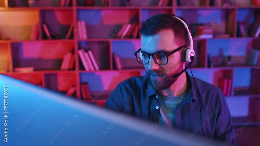 Talking, having online meeting. Man in eyewear and headphones is sitting and working by computer. Neon lighting. Talking, having online meeting. Man in eyewear and headphones is sitting and working by computer. Neon lighting.