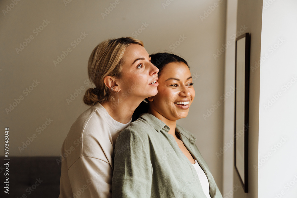 Lesbian couple hugging and looking through window while standing together at home Lesbian couple hugging and looking through window while standing together at home